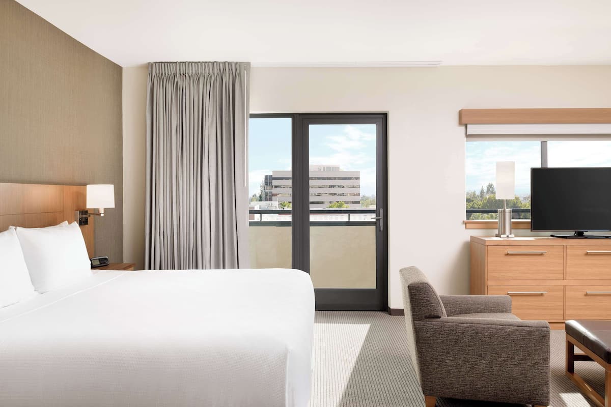 A hotel room with a large bed, a chair, a TV, and a view of a building through the window.