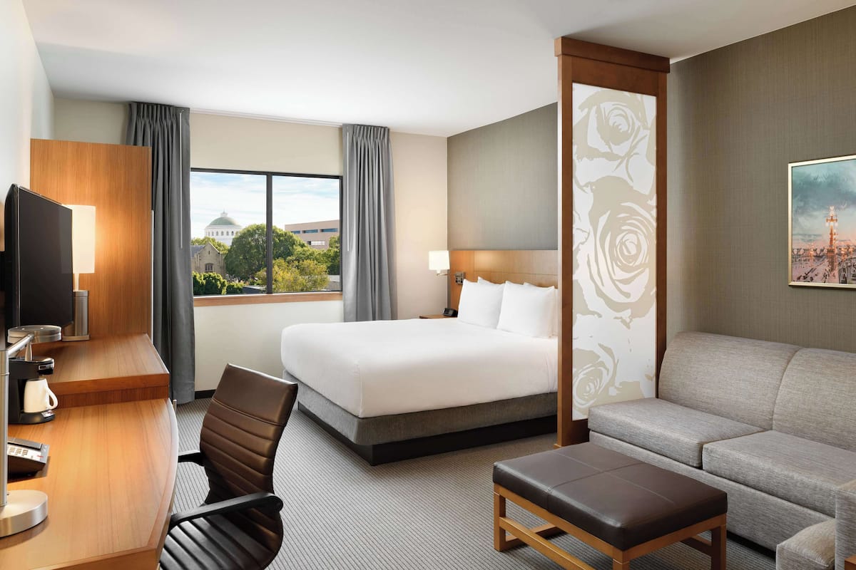 A hotel room with a bed, a desk with a computer, a chair, a sofa, and a window with a view of a building and trees.