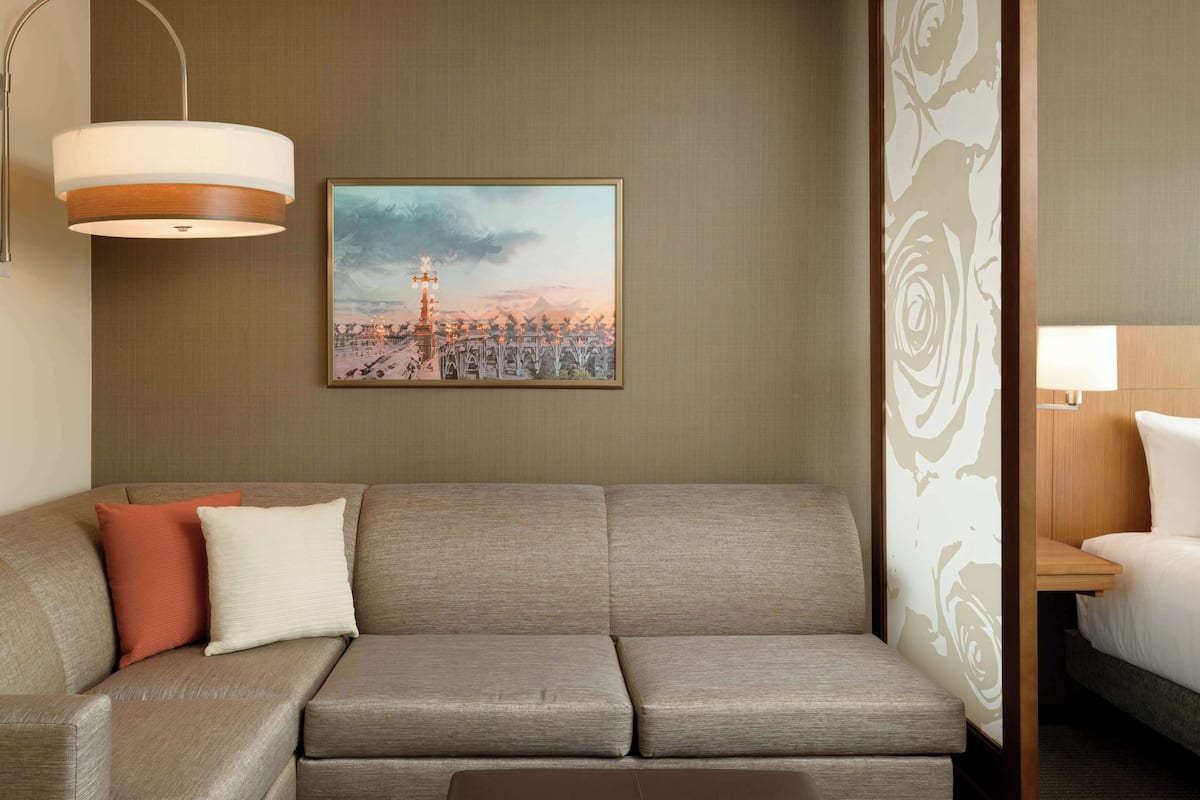 A hotel room with a grey sofa, a framed cityscape painting, a wooden nightstand, and a bed with white bedding.