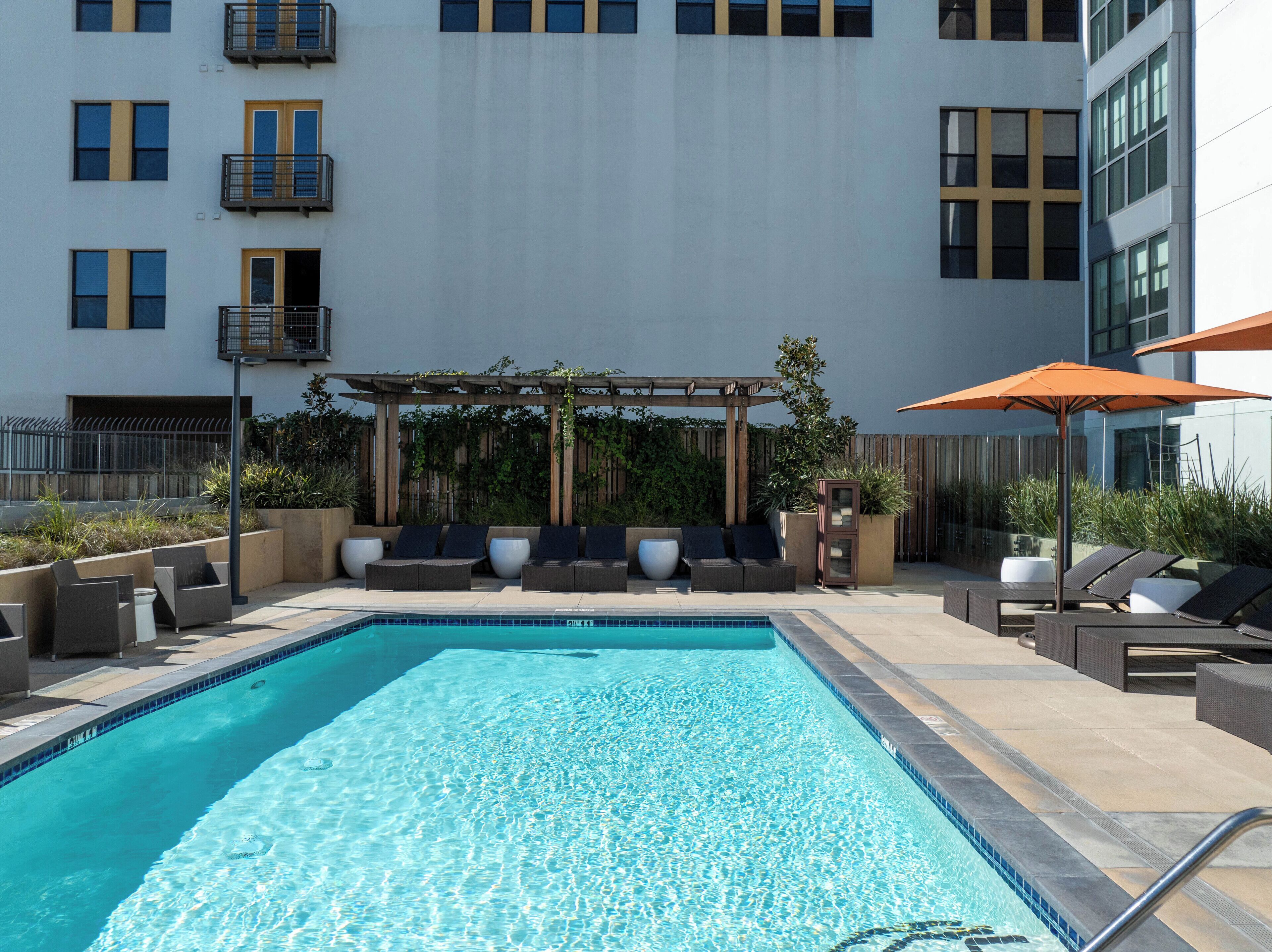 A swimming pool with lounge chairs and umbrellas, surrounded by a building with balconies and a pergola.