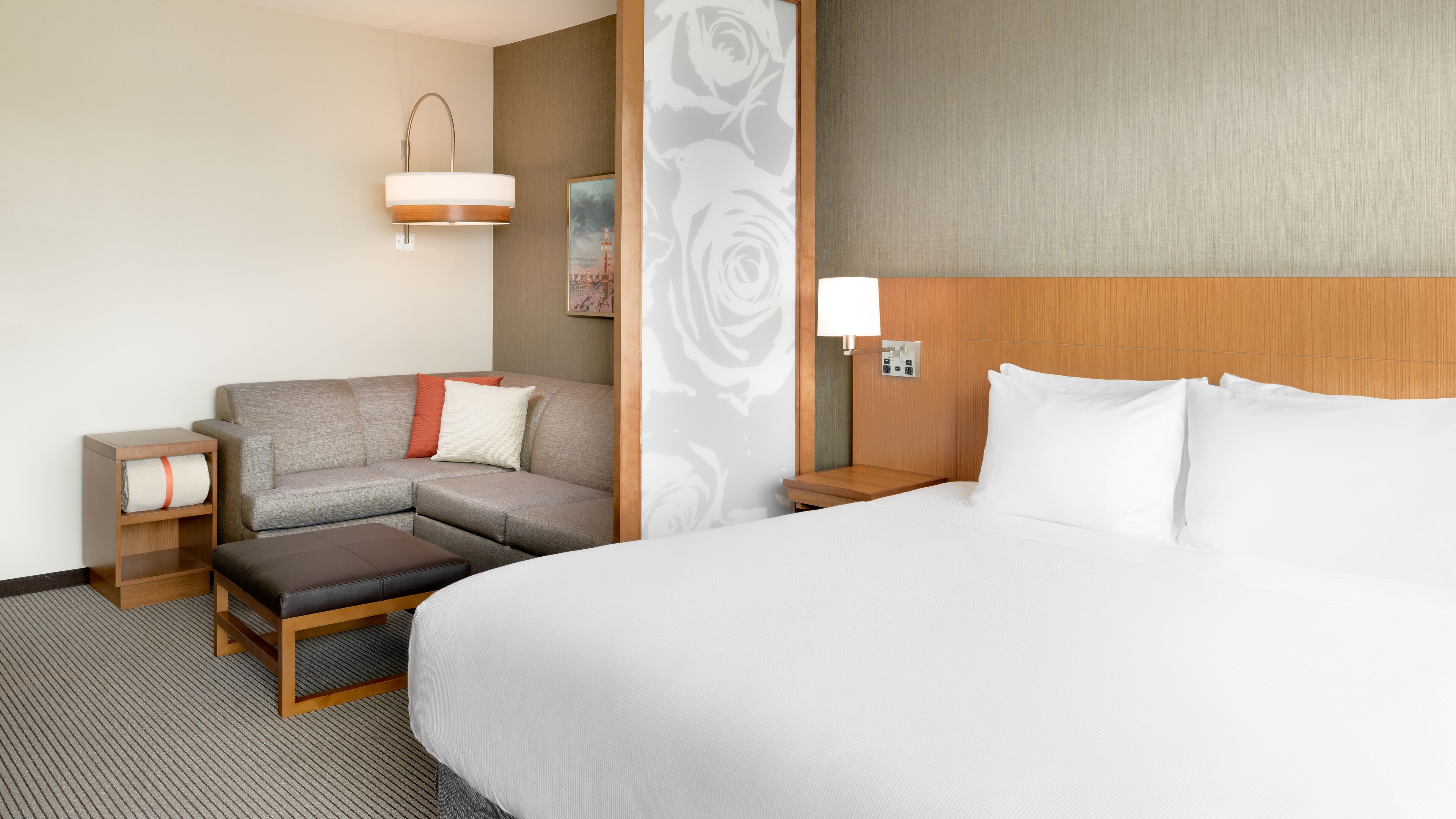 A hotel room with a large bed, a sofa, a small table, a nightstand, and a wall-mounted lamp.