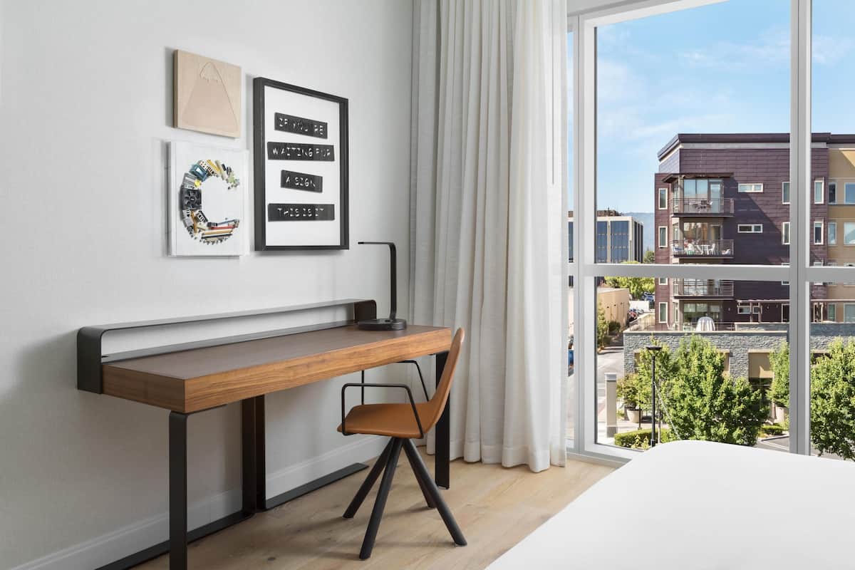 A modern bedroom with a wooden desk, a chair, a bed, and a large window with a view of buildings.