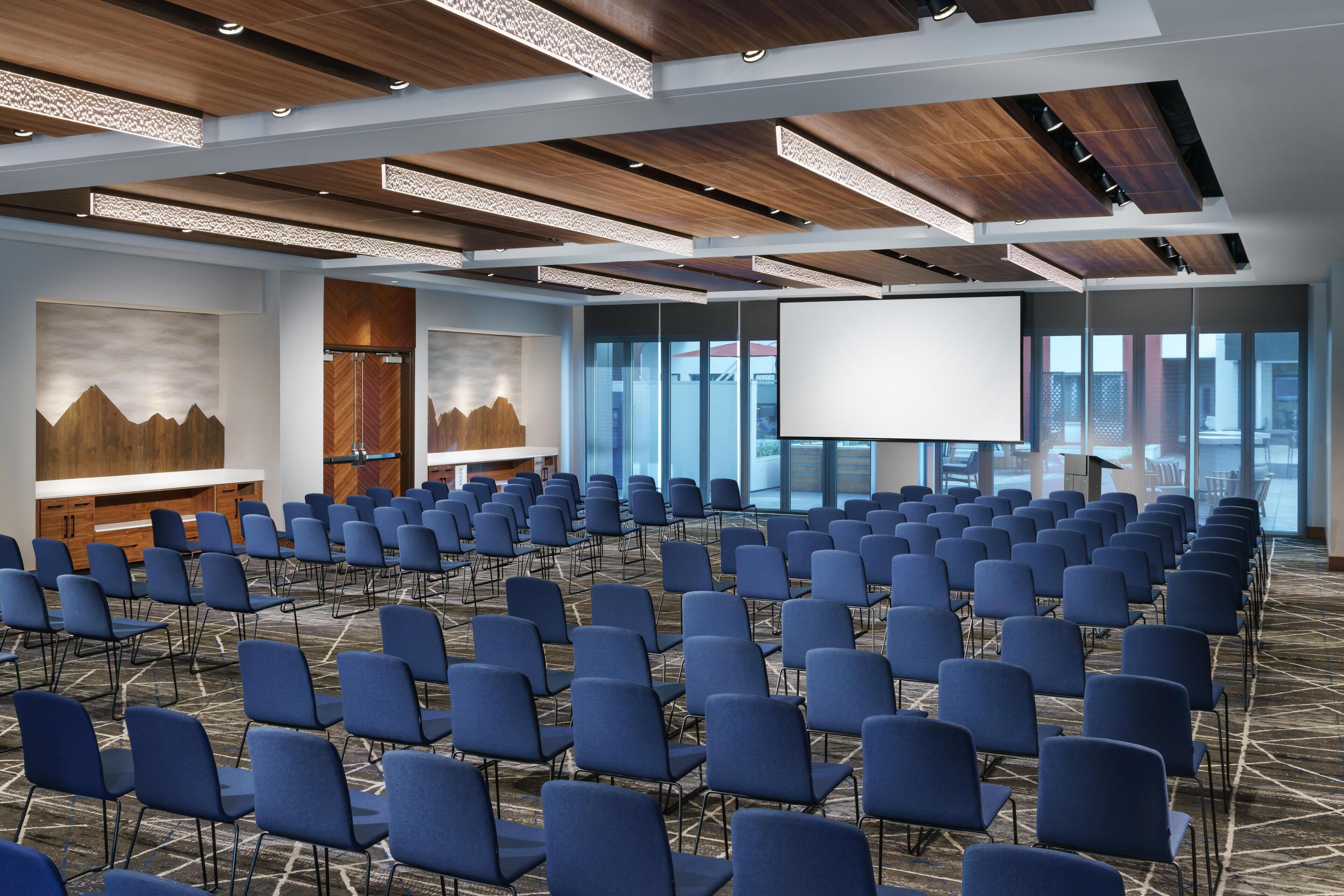 Une salle de conférence avec des rangées de chaises, un grand écran et des panneaux de plafond en bois.