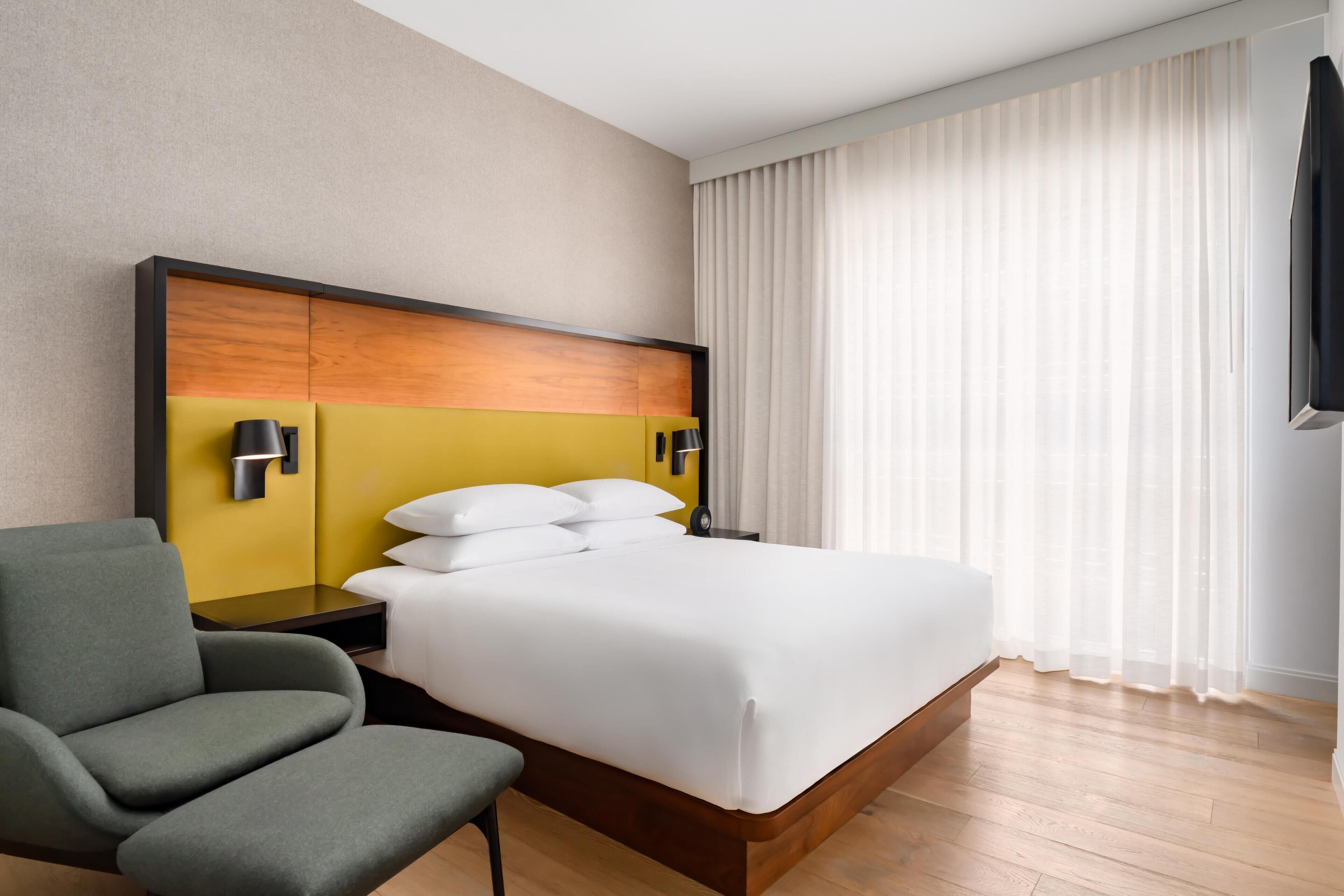 A hotel room with a bed, a chair, a nightstand, a TV, and a window with curtains.