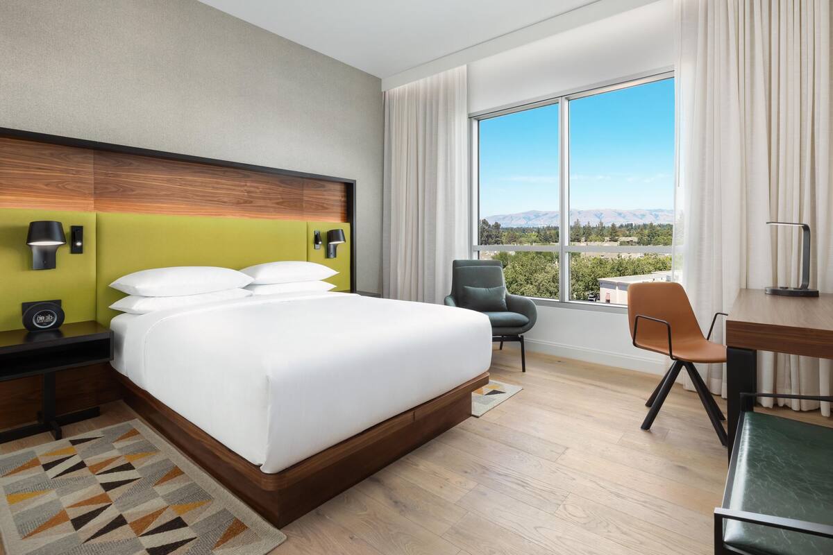 A hotel room with a large bed, a desk, a chair, and a view of a cityscape through a large window.