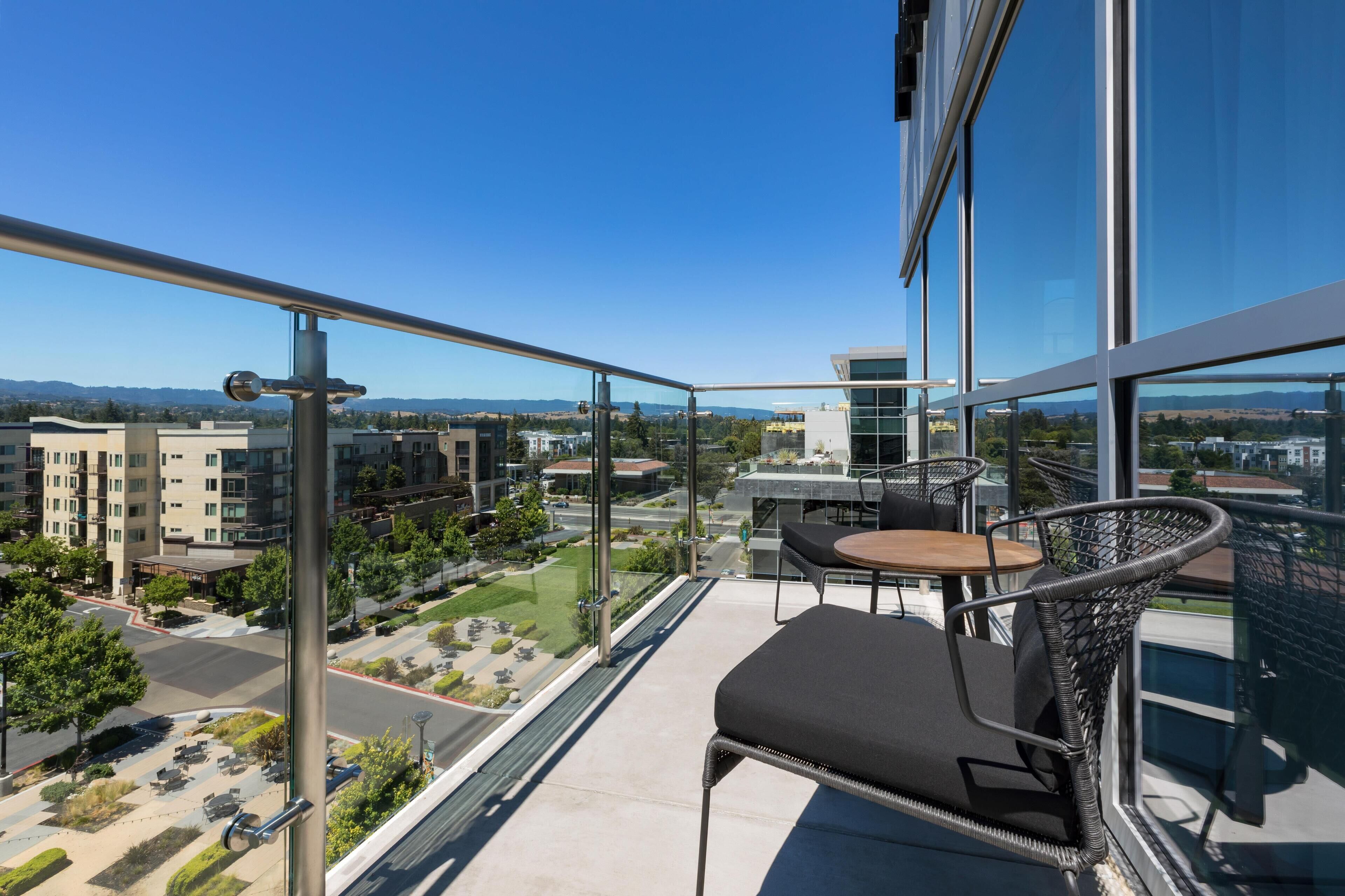 A balcony with a glass railing, a wicker chair, a small table, and a view of a cityscape with buildings and greenery.