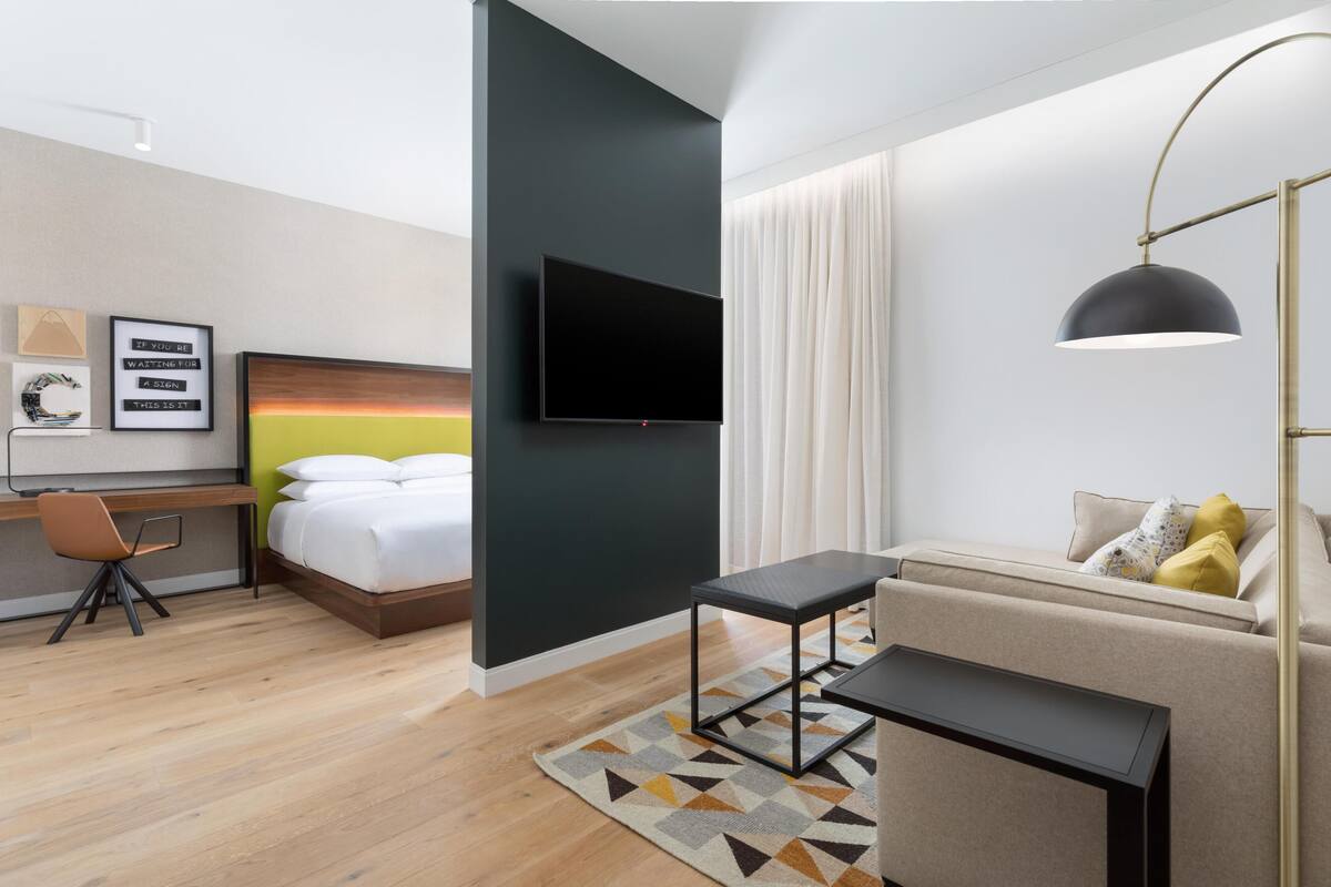 A modern hotel room with a bed, desk, TV, sofa, and a desk lamp.