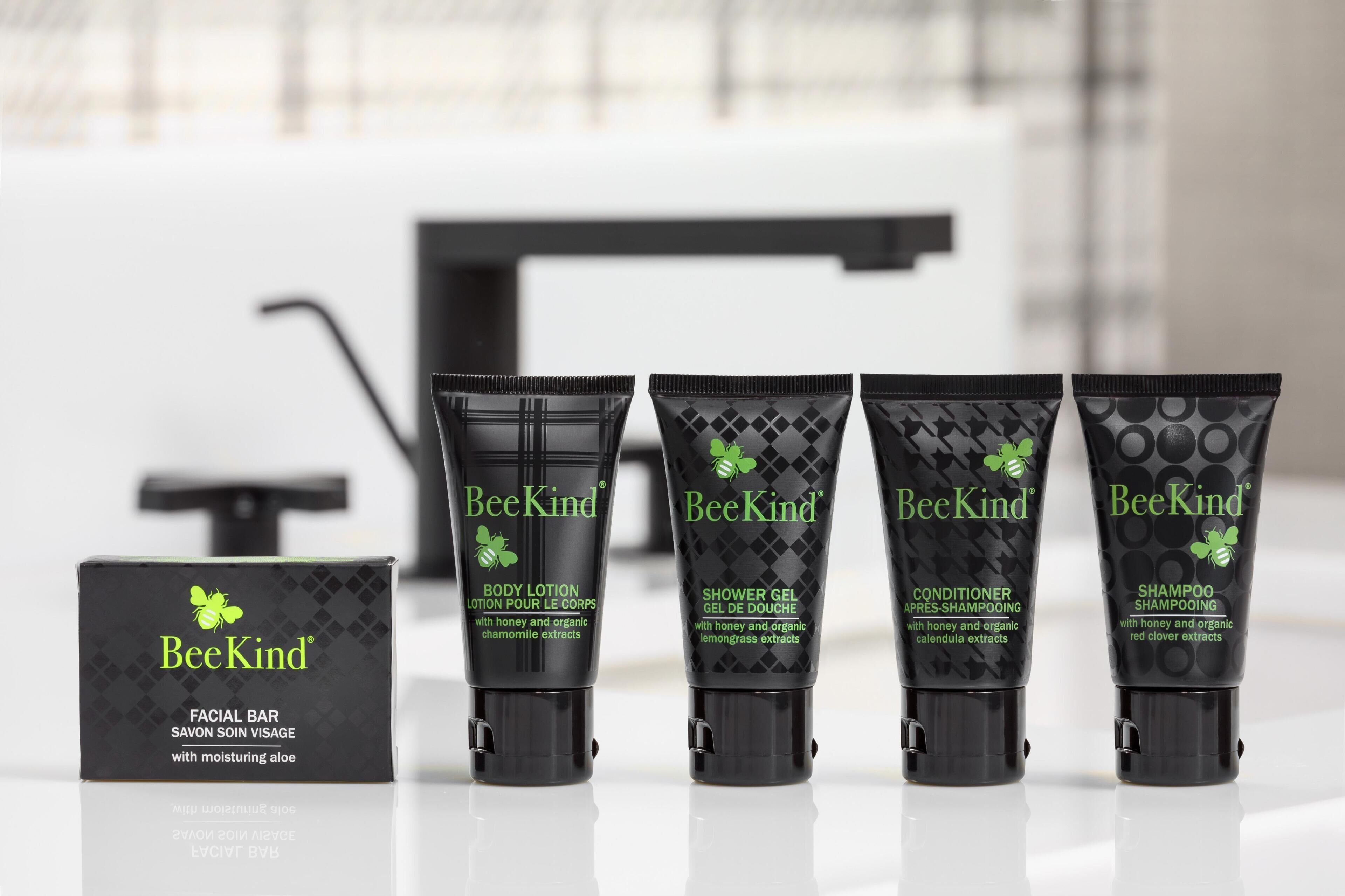 A BeeKind facial bar, body lotion, shower gel, conditioner, and shampoo arranged on a reflective surface.