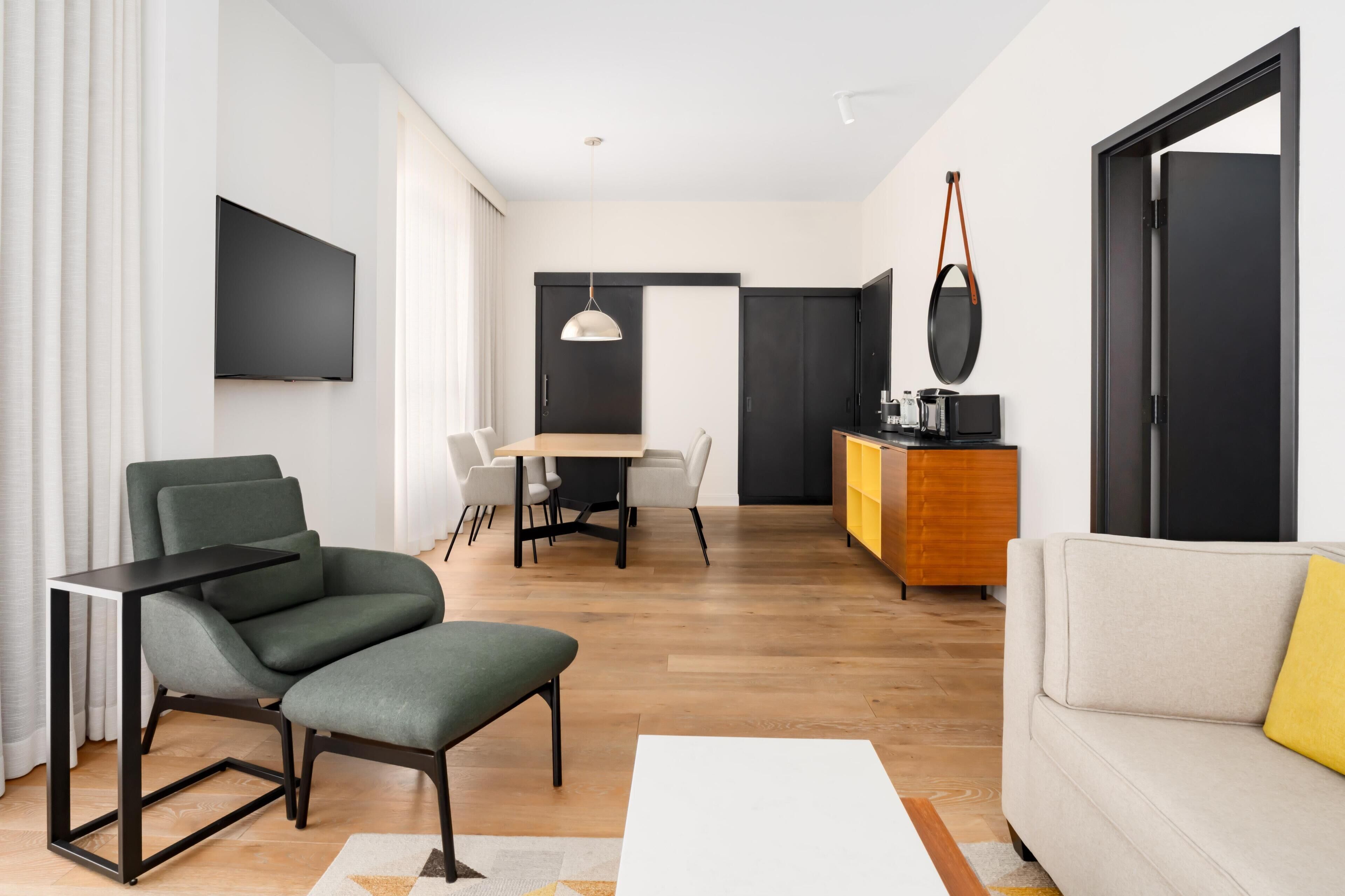 A modern living room with a sofa, a dining table with chairs, a TV mounted on the wall, and a kitchen area with a counter and appliances.