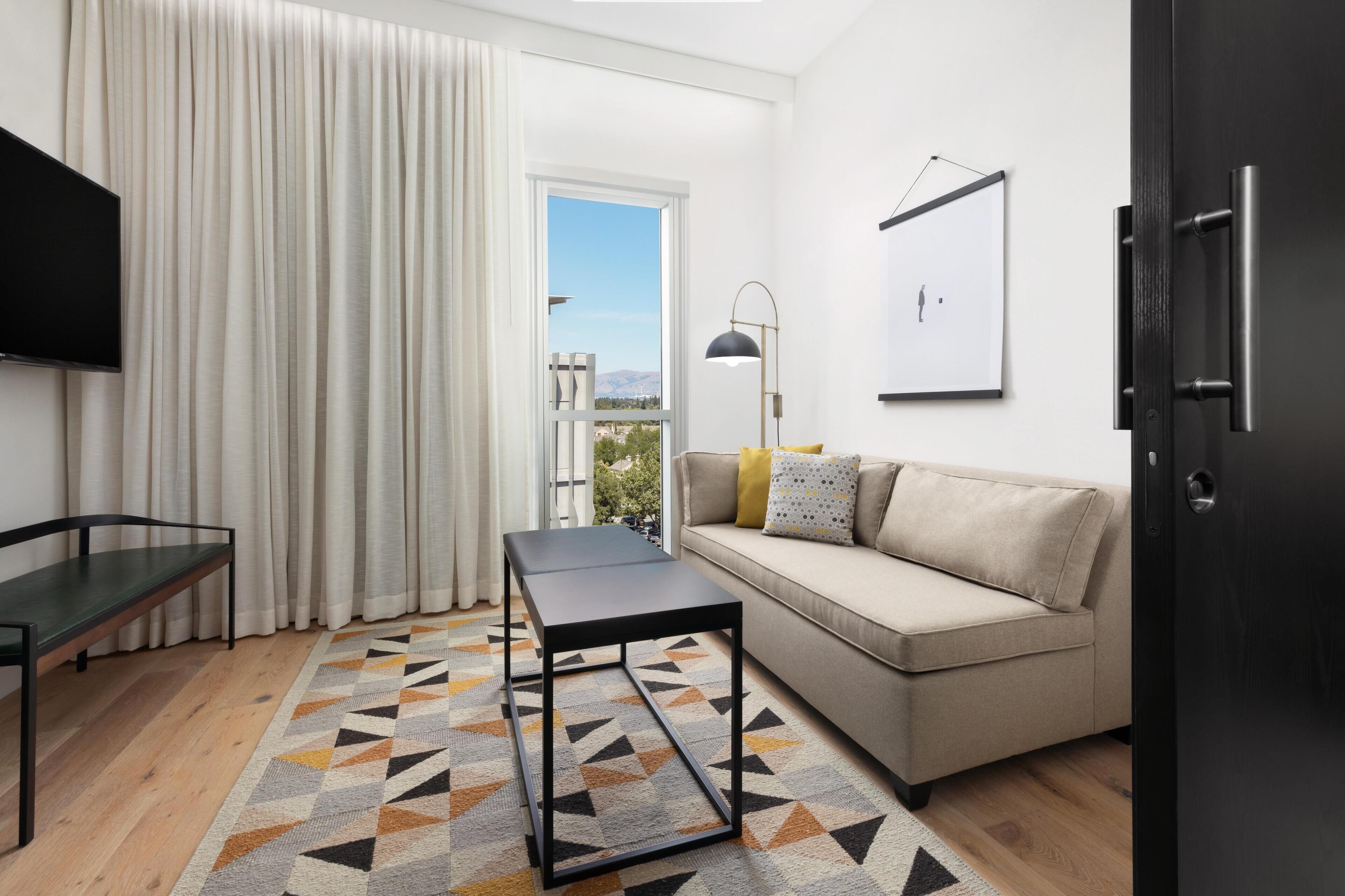A modern living room with a sofa, a coffee table, a TV, and a large window with curtains.
