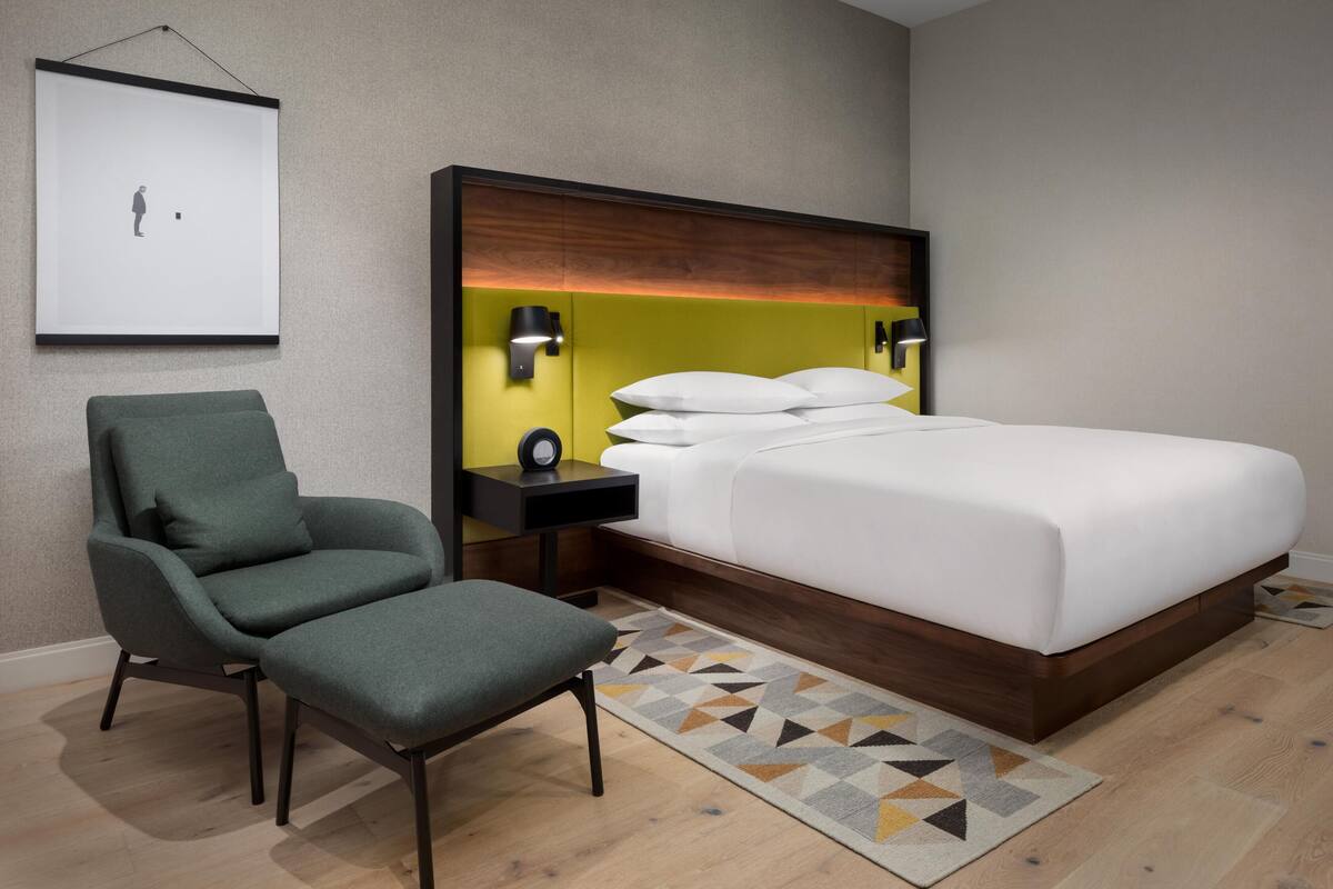 A hotel room with a bed, a chair, a nightstand, a wall-mounted projector, and a patterned rug.