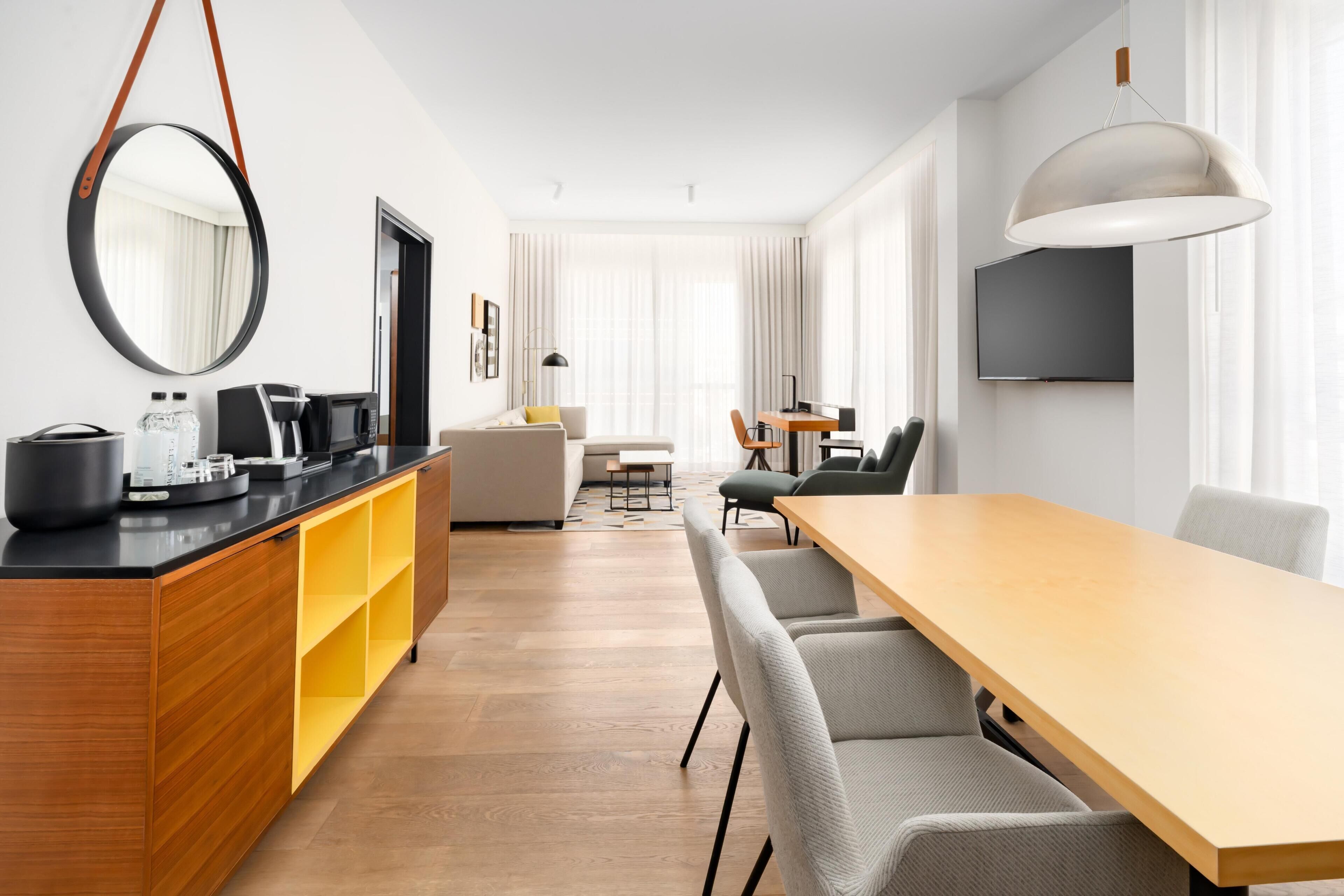 A modern hotel room with a wooden desk, a large mirror, a flat-screen TV, a sofa, a dining table, and a large window.
