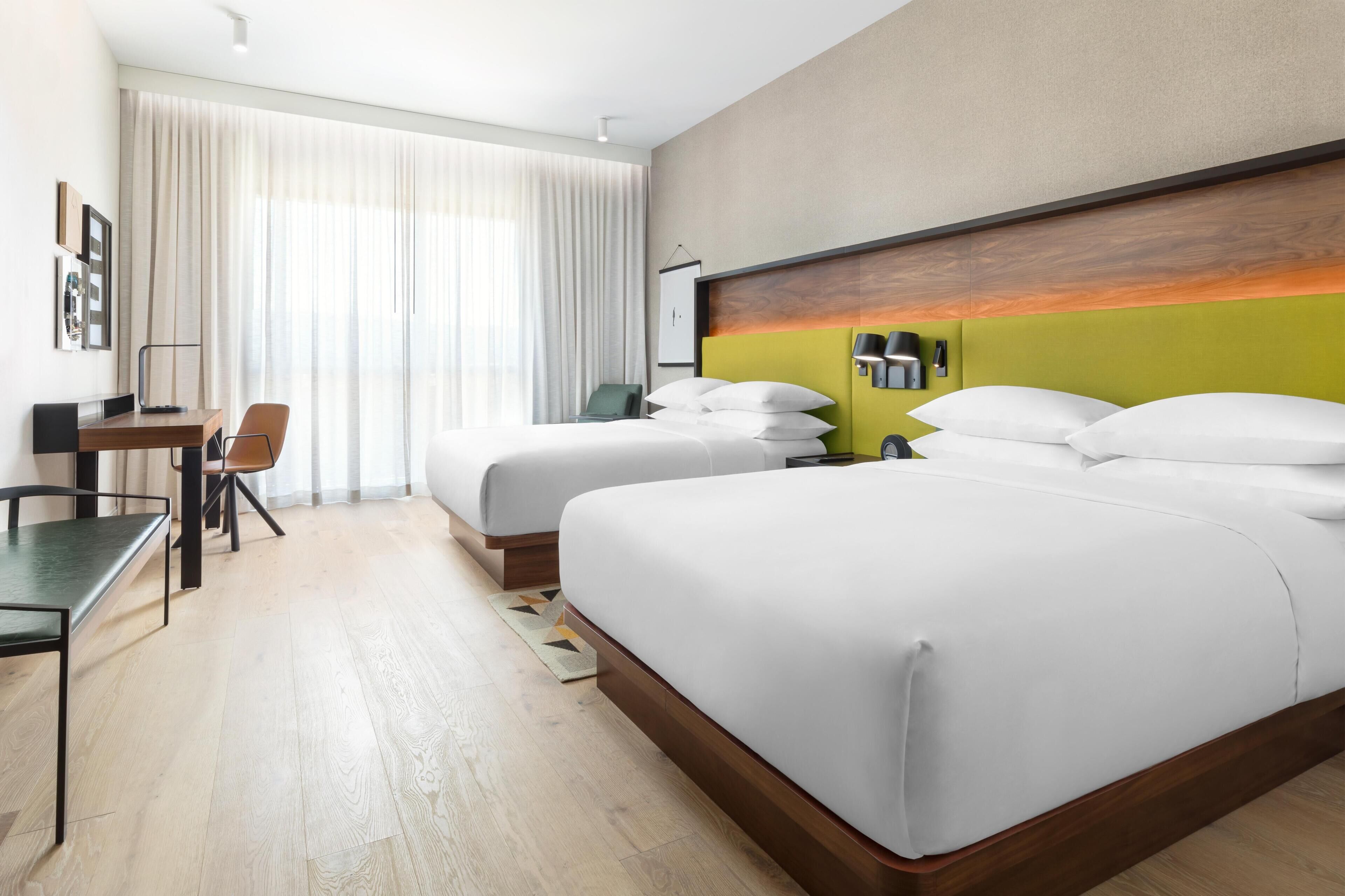 A hotel room with two beds, a desk, a chair, and a large window with curtains.