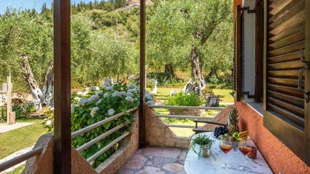 A patio with a table set for two, surrounded by a stone wall and a railing, overlooking a garden with trees and flowers. - Anneta Studios (Igoumenitsa)