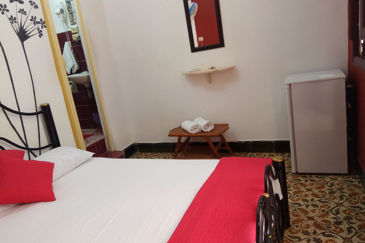 A hotel room with a bed, a small wooden table with towels, a mirror, and a refrigerator.