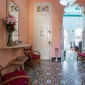 Une pièce avec un sol à motifs, une porte blanche, un miroir, un vase de fleurs et des meubles en osier.