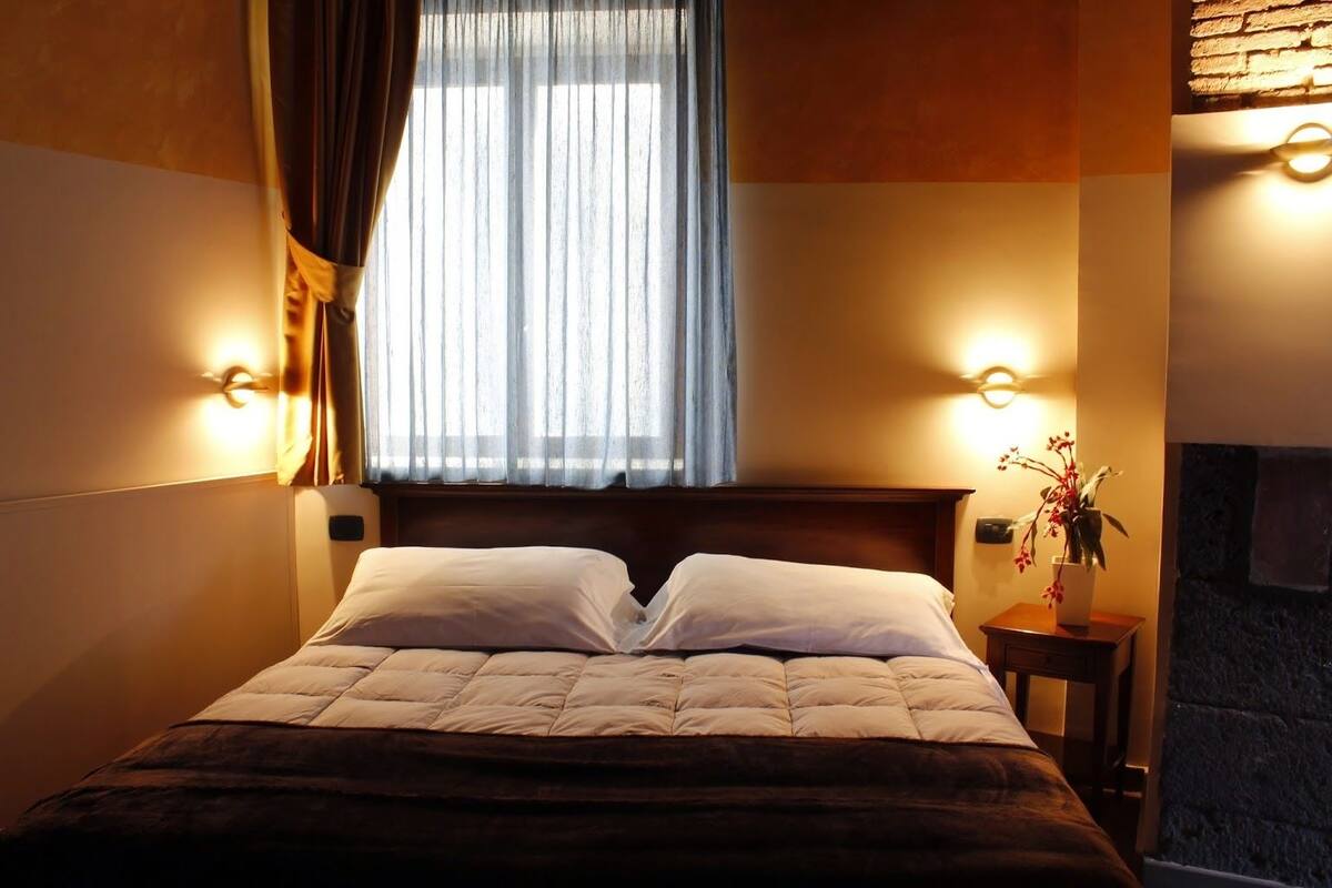 A neatly made bed with a headboard, two wall-mounted lights, a window with curtains, and a small table with a vase of flowers.