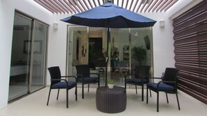 Et udendørs opholdsområde med et bord, stole, en blå parasol og en glasdør, der fører til et indre rum.
