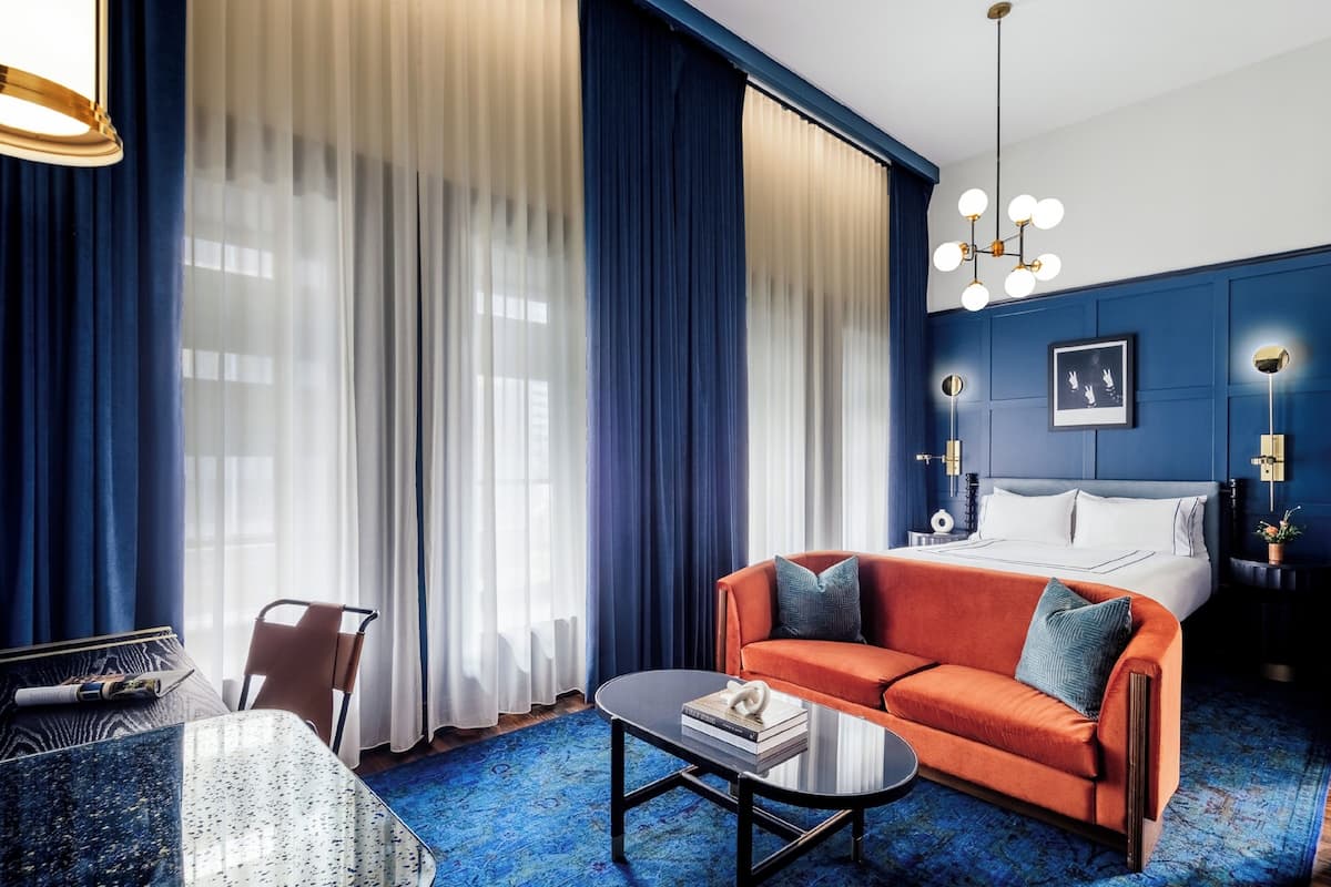 A modern hotel room with a blue and white color scheme, featuring a sofa, a bed, a coffee table, and a dining area.