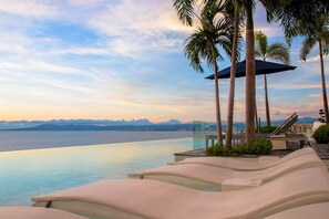 Une piscine à débordement avec des chaises longues, des palmiers et une vue dégagée sur la mer et les montagnes lointaines.