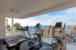 Une salle de sport équipée de tapis de course et de vélos d’exercice, avec de grandes fenêtres offrant une vue sur la ville.