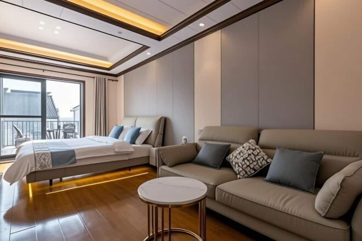 A modern hotel room with a bed, a sofa, a small round table, and a large window offering a view of the outside.