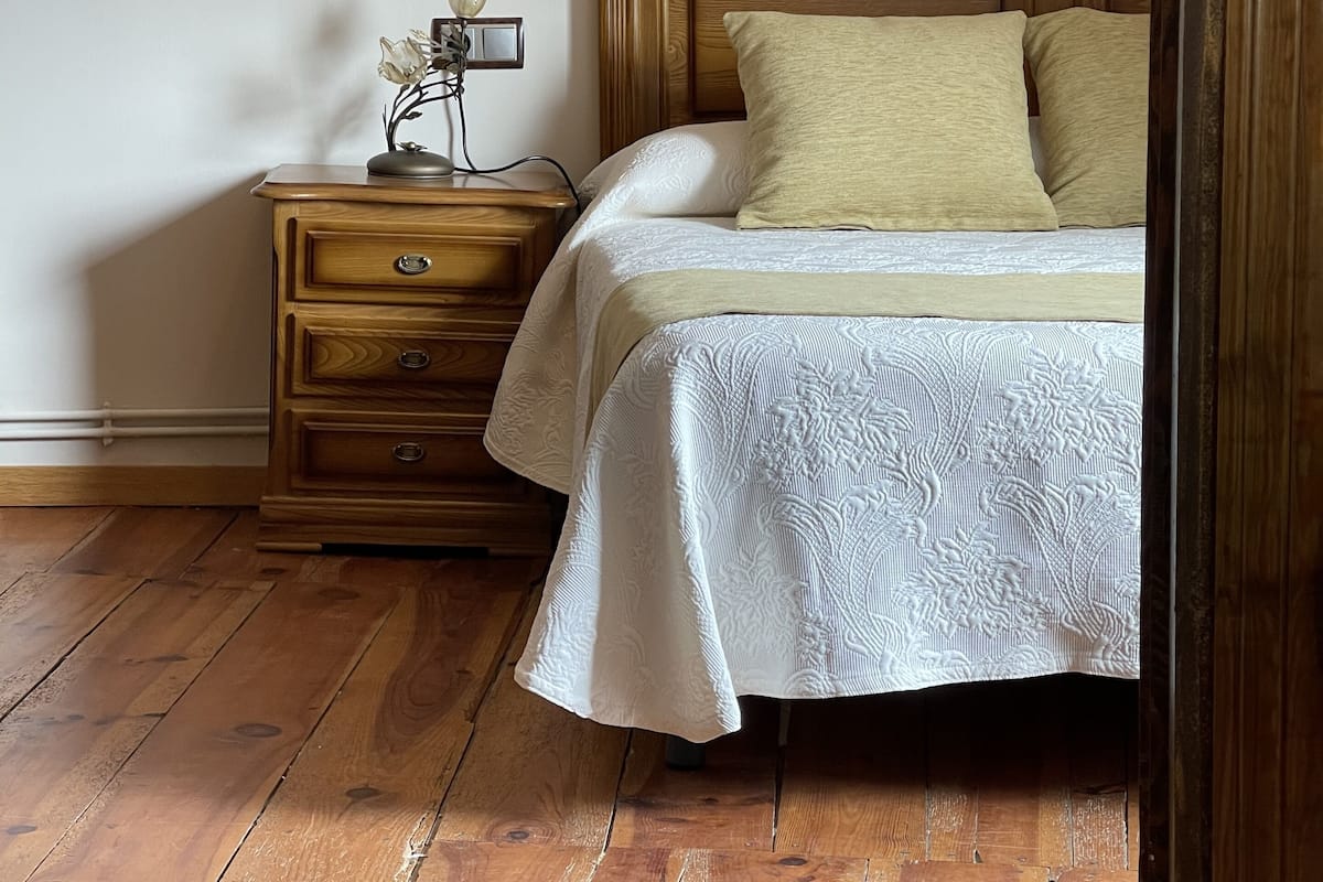 A bedroom with a wooden headboard, a wooden nightstand with drawers, a bedside table with a lamp, and a patterned bedspread.