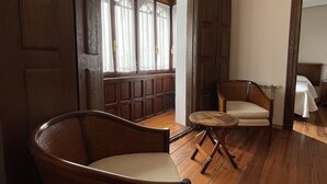 Una habitación con suelo de madera, una silla de mimbre, una pequeña mesa redonda y un banco con cortinas.