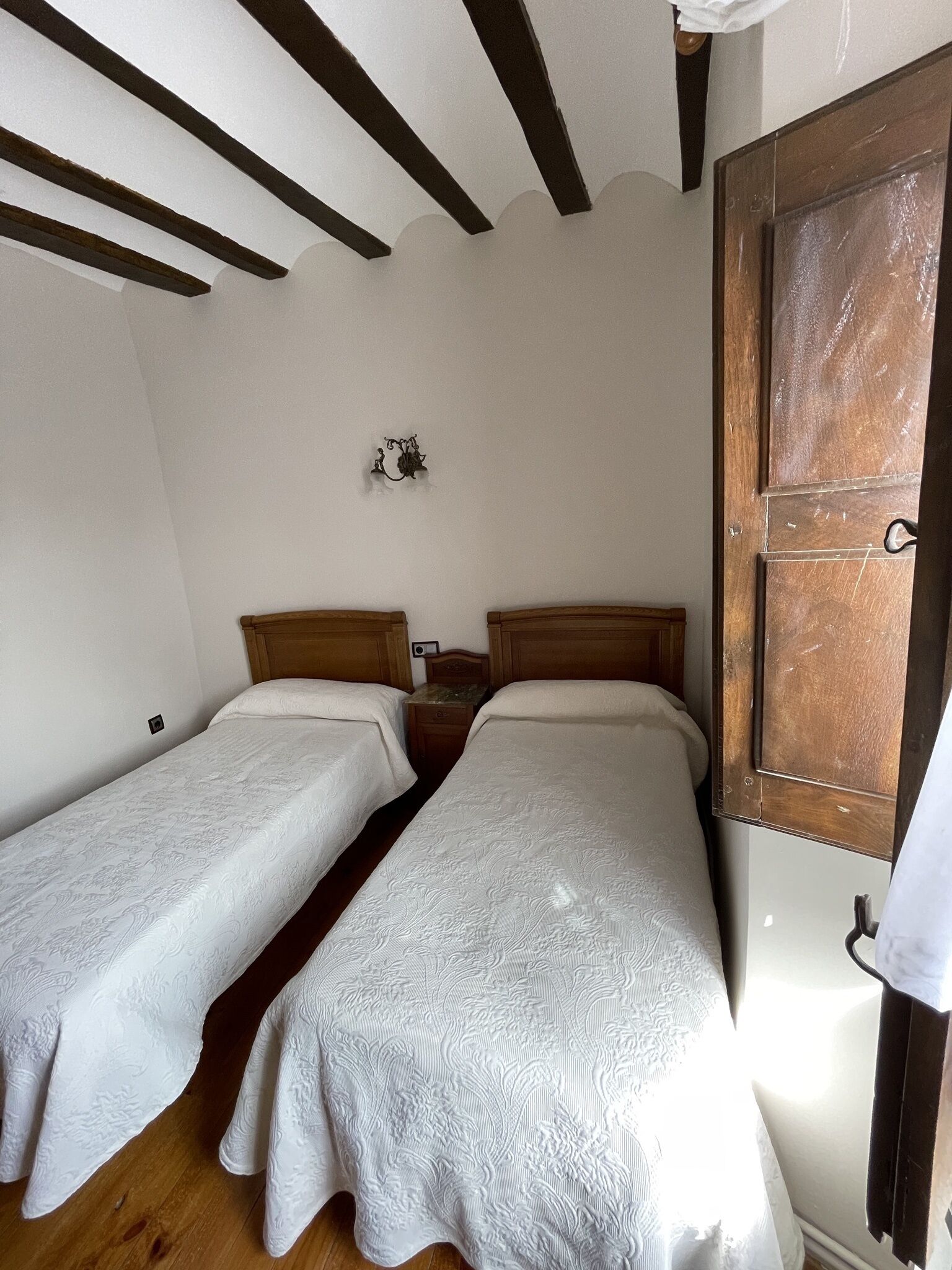 Una habitación con dos camas individuales, techo de madera, una lámpara pequeña en la pared y una puerta de madera.