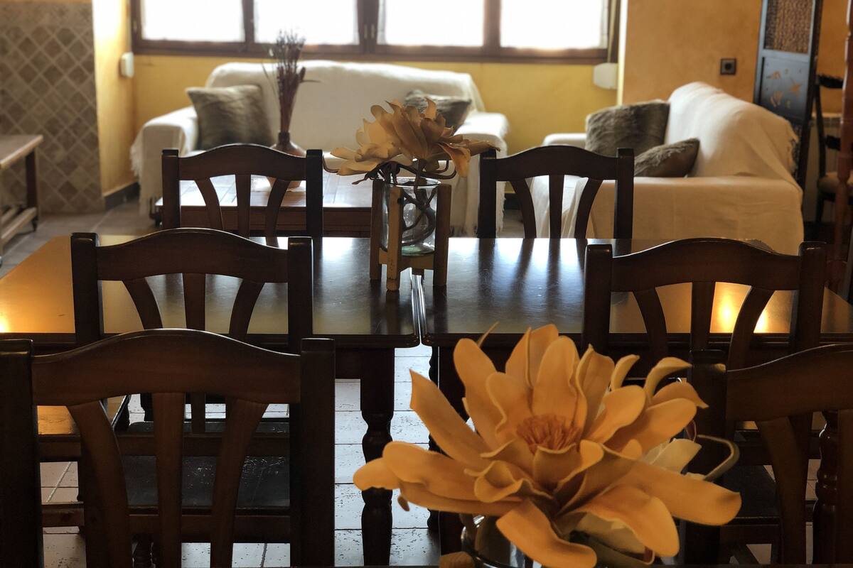 A dining area with a wooden table and chairs, a vase with a flower arrangement, and a view of a living room with a sofa and cushions in the background.