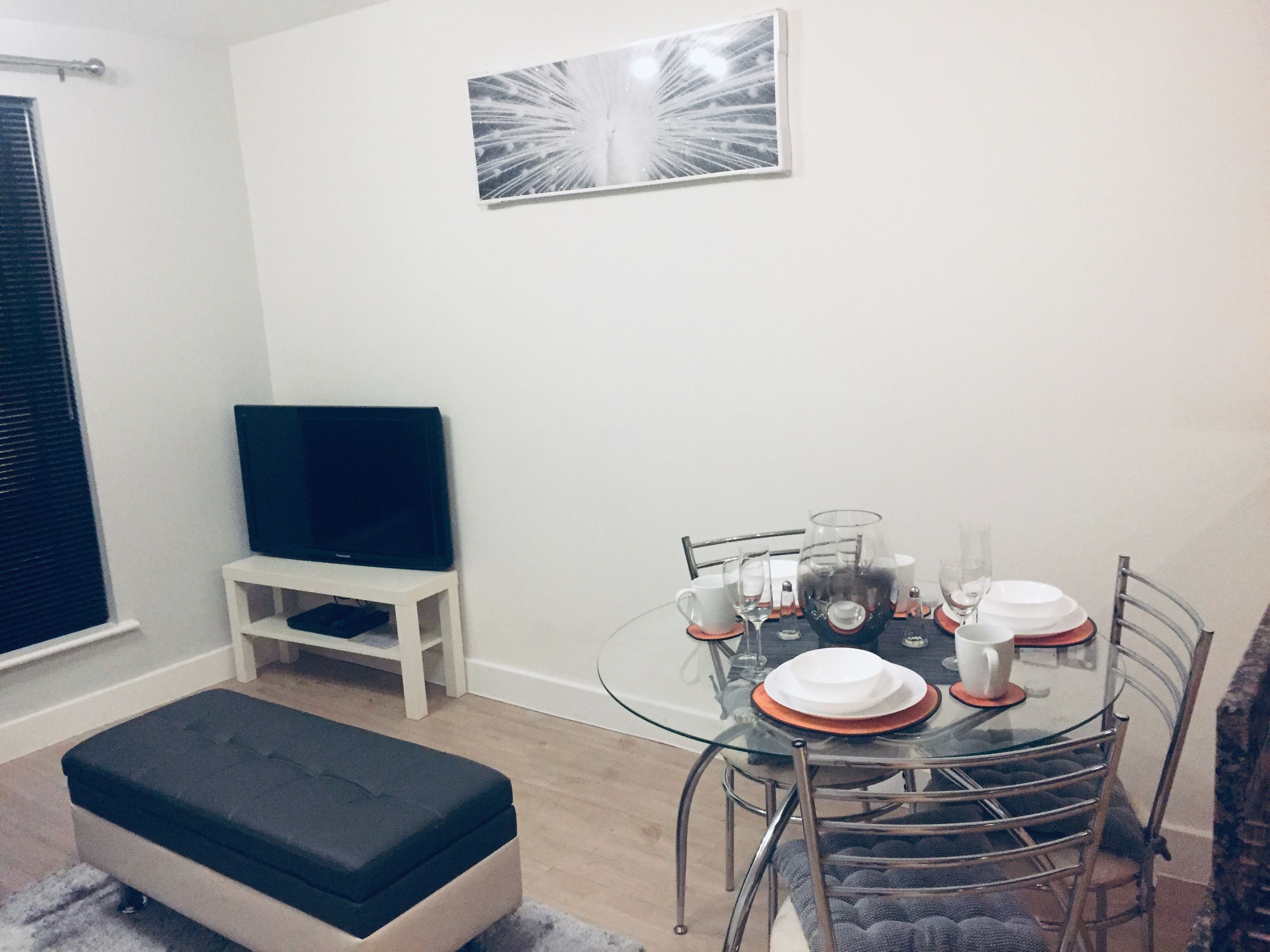 A dining area with a glass table set for four, surrounded by metal chairs. A white bench is against the wall, and a flat-screen TV is mounted above it.