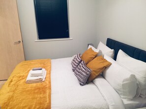 A neatly made bed with white and brown bedding, a window with blinds, and a wooden door. - The Gallery (Nottingham)