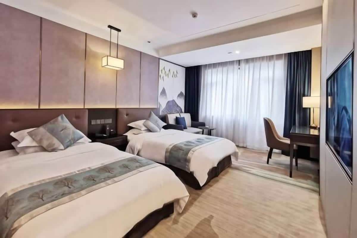 A hotel room with two beds, a desk, a chair, a TV, and a large window with curtains.