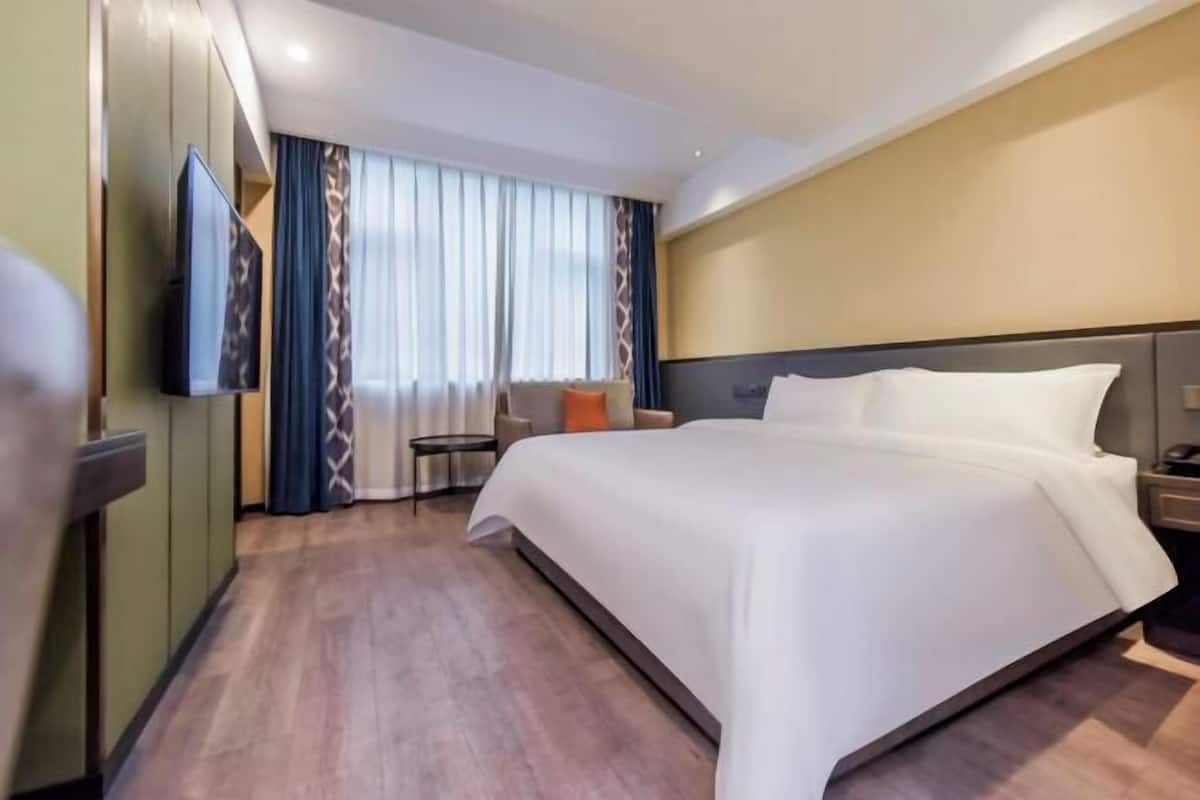 A hotel room with a large bed, a flat-screen TV mounted on the wall, a seating area with a chair and a small table, and a large window with curtains.