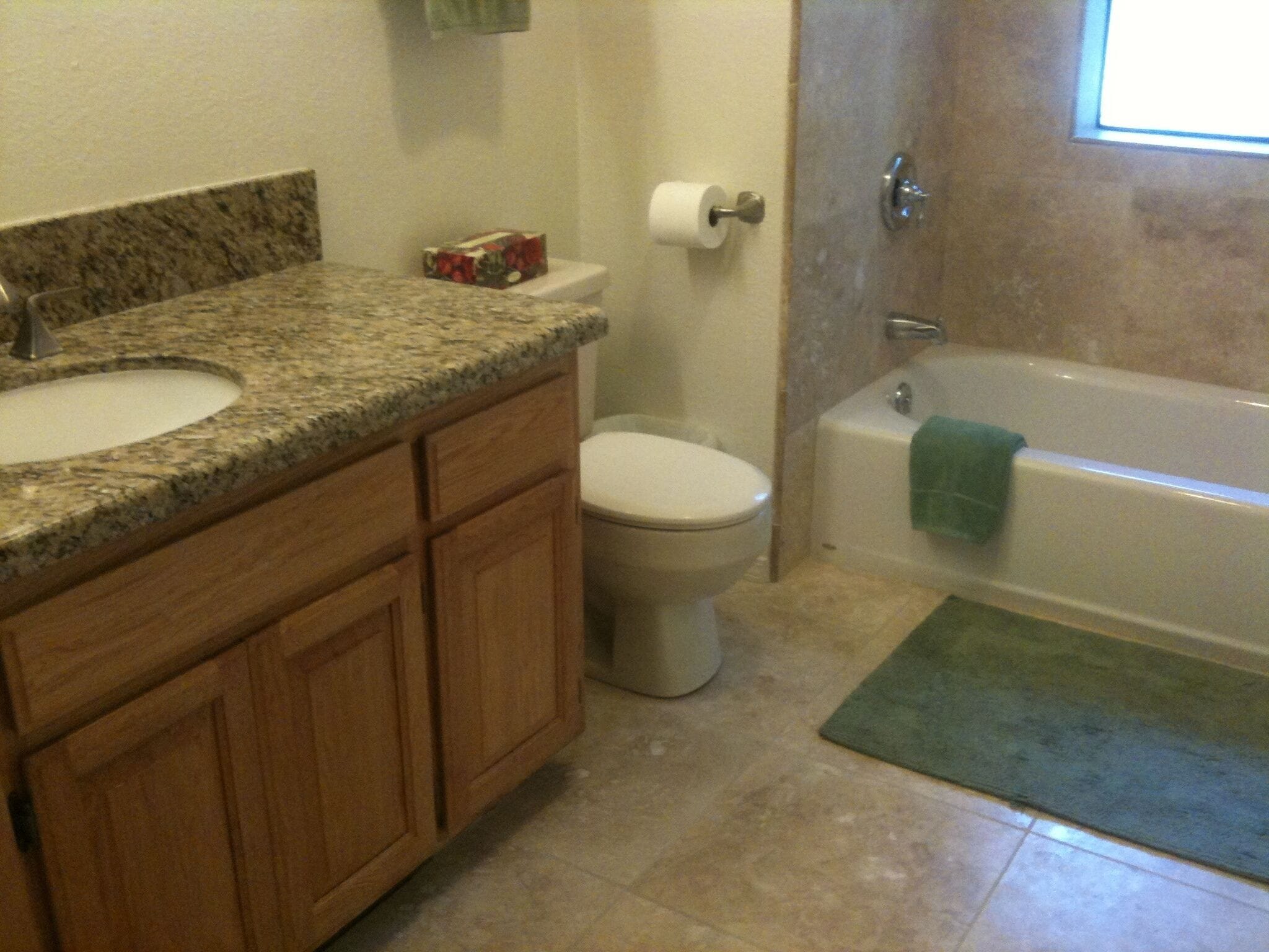 A bathroom with a granite countertop sink, wooden cabinets, a toilet, and a bathtub with a green towel.