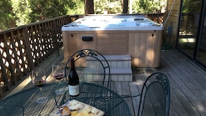 Une terrasse en bois avec un jacuzzi, une table sur laquelle se trouvent une bouteille de vin, du fromage, des crackers et un verre de vin.