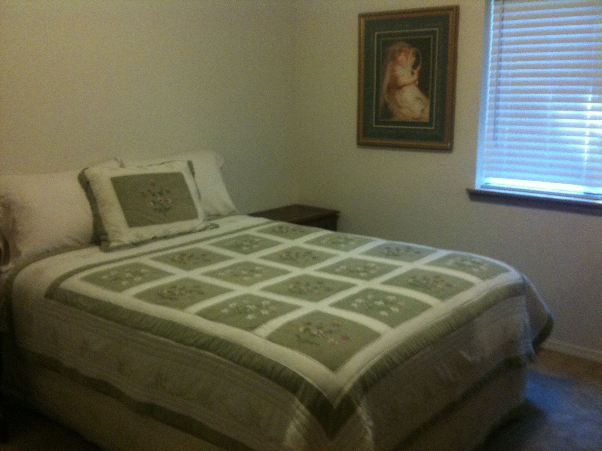 A bedroom with a bed, a nightstand, a framed picture on the wall, and a window with blinds.