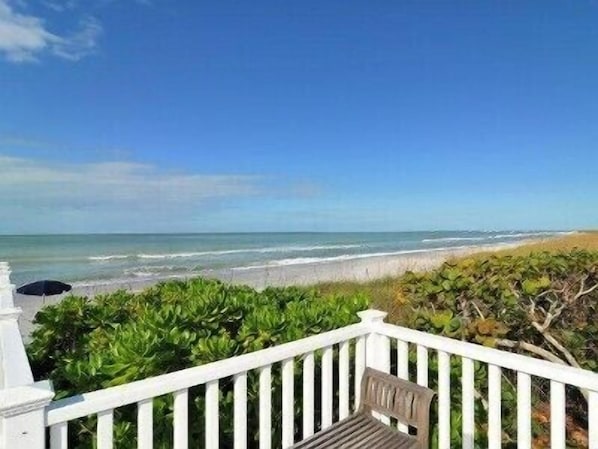 A beach view from a balcony with a white railing, a bench, and a view of the ocean.