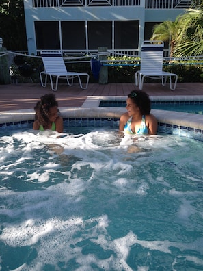 A swimming pool with two people sitting in the water. There are lounge chairs and a building in the background. - Escape to Paradise – Beautiful & Budget-Friendly Condo! (Rum Point)