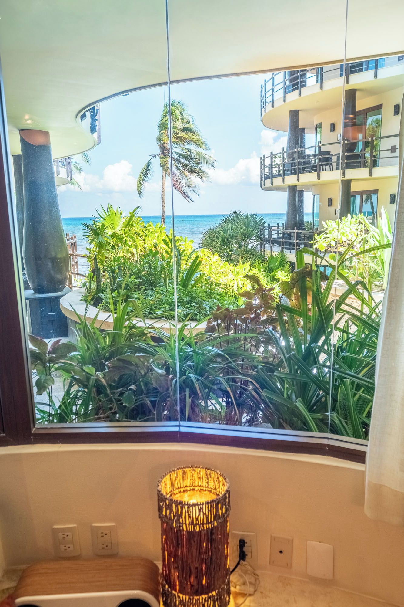 A room with a view of a pool and palm trees, a wicker lamp, and a wooden bench.