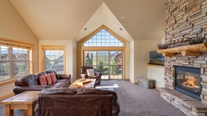 A spacious living room with a stone fireplace, leather sofas, a wooden coffee table, and large windows offering a view of the surrounding landscape. - 67 Fireweed | 4BR Hot Tub, Sleeps 13 (Tabernash)
