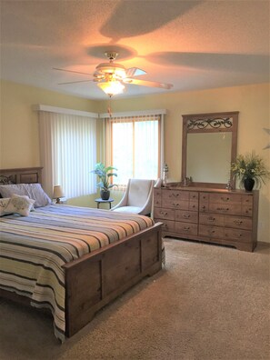 A bedroom with a large bed, a dresser, a chair, a ceiling fan, and a window with curtains.