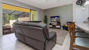 Ein modernes Wohnzimmer mit grauem Ledersofa, einem Essbereich mit Holzsesseln, einem an der Wand befestigten Flachbildfernseher und einer Glastür, die auf einen Balkon führt.