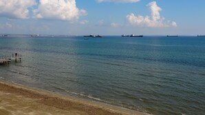 A coastal scene with a pier extending into calm waters, several large cargo ships, and a clear sky with scattered clouds.