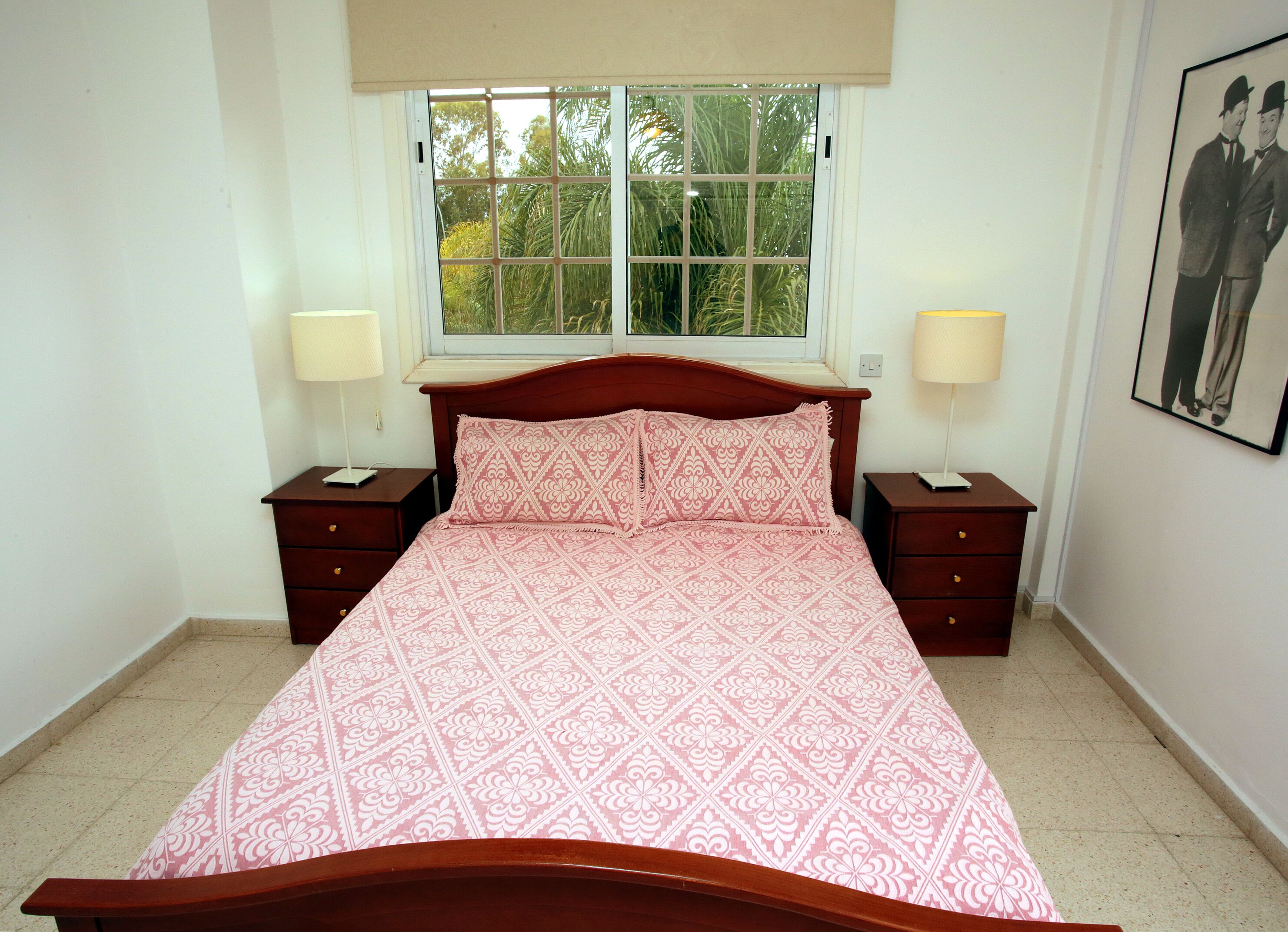 Un dormitorio con una cama de madera, dos mesitas de noche, una ventana con vistas a árboles y un cuadro en la pared.