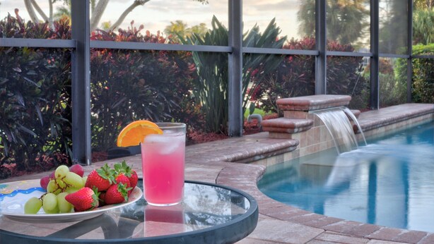 Um copo de bebida rosa com morango enfeitando e um prato de frutas sobre uma mesa, perto de uma piscina com cachoeira.