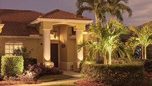 Uma casa com telhado telhado, varanda coberta com colunas, e um jardim bem cuidado com palmeiras e arbustos.