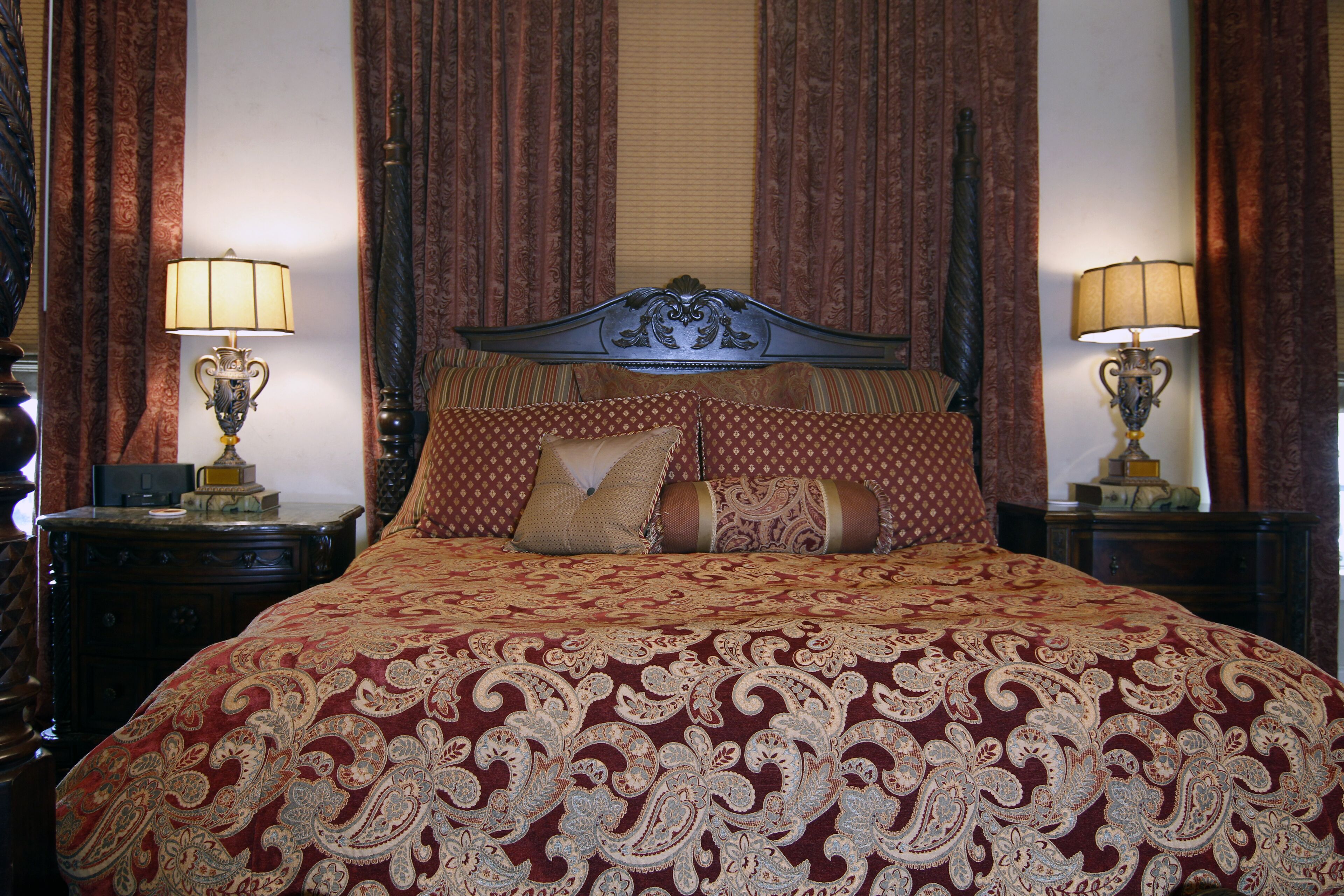 A large bed with a patterned bedspread, two bedside tables with lamps, and heavy drapery.