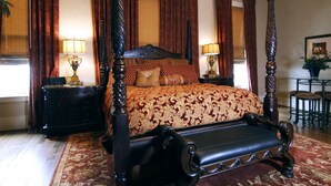 A four-poster bed with a dark wooden frame and a red and gold patterned bedspread. There are two nightstands with lamps on either side of the bed. The room has a chandelier and a red carpet. - Legacy Suite on Main St in central downtown, jacuzzi tub, ask about Wine Tours!! (Fredericksburg)