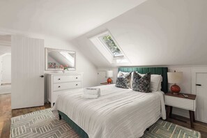 A bedroom with a sloped ceiling, a bed with a white comforter, a nightstand with a lamp, and a dresser with a mirror.